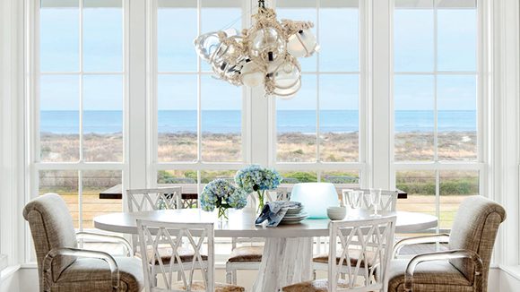 A floor-to-ceiling window frames the view next to a round, light-colored dining table surrounded by white wooden chairs with geometric wood carved back detail. Head chairs are a darker, textured tan with clear Perspex arms. The chandelier is made from glass balls and floats tangled in nets.
