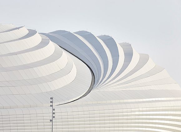 Image Al Wakrah Stadium courtesy Dezeen, the building is a series of stepped surfaces clad in white that cast light shadows like a ripple.