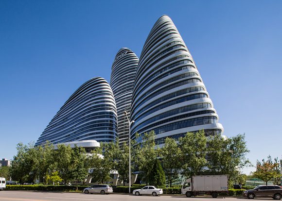 The Wangjing Soho building looks like 3 tall pebbles balanced against each other to stand, with bold lines of windows looking like layers of rock or sediment. The building is surrounded by greenery at the bottom, and a blue sky above.