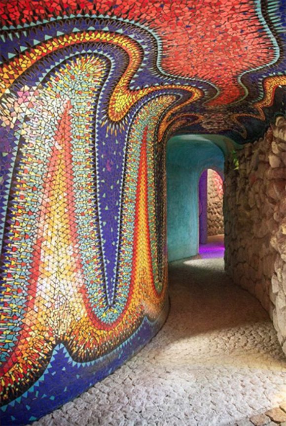 Image Nido De Quetzalcoatl courtesy @javiersenosiaina on Instagram. A left wall in a stone tunnel of sparkling mosaic pieces form tall waves or flickering candles, with a sun in the middle near the ceiling. The colors are mostly midnight blue, with sky blue, orange, yellow, gold, and black. The stone is a light tan color.