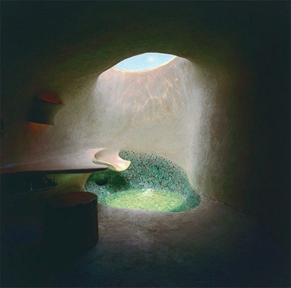 Image Casa Orgánica-Ducha courtesy @javiersenosiaina on Instagram. The photo is of a cave, with a hole in the center of the ceiling where light pours in, and strikes a glass mosaic pool of mostly green with blue and black accents. An off-white shape to the left provides water through its tube-with-a-mouth-like shape. The rest of the space is veiled in darkness.