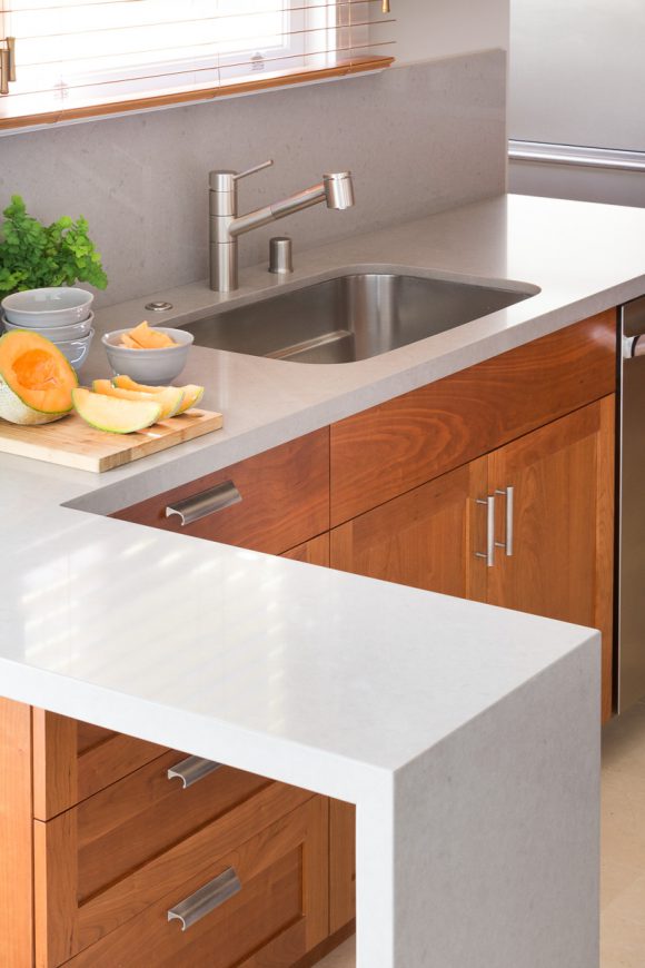 A close-up view of an accessible kitchen featuring cherry wood cabinet doors and drawers with easy-pull handles, a grey countertop that fits a walker or wheelchair underneath the L-shaped end of the counter, and an easy-to-use sink faucet fixture in silver. The counter is dressed with sliced melon and bowls, and a sprig of green plant next to the sink.