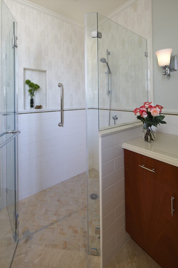 A modern bathroom featuring a curbless walk-in shower surrounded by clear glass, with a silver metal grab bar on the wall next to a niche dressed with green plant and loufah. The far wall shows a hand-held shower fixture in silver against hexagonal grey and white toned tiling. Below a line of horizontal dark grey tiles, the bottom half is plain white subway tiling in a medium-size scale, and the flooring is a warm beige and tan toned porcelain, laid at an angle to make the space feel more open. To the right of the shower is a corner of the dark wood cabinetry with silver metal drawer and door pulls, and a beige countertop, dressed with a vase of orangey-white roses.