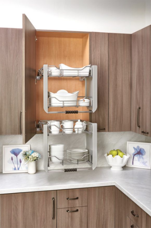 Photo of a modern kitchen with light vertically-striated wood cabinets, bronze metal pull handles, a white marble countertop. One cabinet is open to reveal a pull-down metal shelving unit filled with dishes. The inside back of the cabinet is a creamy orange color, like a popsicle, and the countertops are dressed with small framed art of purple flowers, a bowl of green apples, and a vase of fresh flowers in white, green, and light blue.