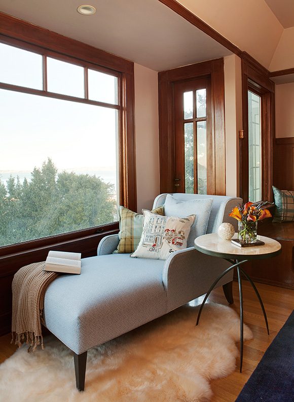 A light-blue custom chaise long with arms is dressed with 3 different patterned and colored pillows, a book, and a textured tan blanket. Next to that is a 3-legged side table dressed with flowers and a small sculptural globe. Both furniture pieces sit atop a fluffy white fur rug, next to a window framed with warm, orangey wood, without any window dressing. Green trees are visible outside the window.