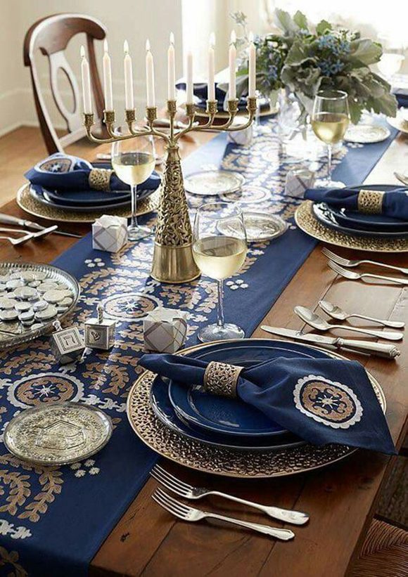 A wooden table is covered with a blue, white and gold Hannukah table runner in the center, with a Menorah sitting in the center, and two dreidels nearby. Glasses of white wine are at each setting. A pair of blue plates sit atop one another over gold plates. Blue napkins with gold and white embroidery that match the tablerunner are weighted by gold napkin holders. Golden tableware sit at each place. A vase of fresh flowers sits at the end of the table, near a window.