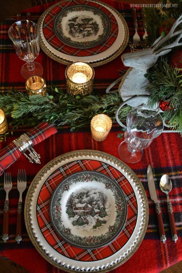 On a beautiful wool blanket of red, black, white and yellow tartan is placed a gold-edged white plate, with a red, black, white and yellow tartan pattern. On top of that is placed a wintry snow scene of a home surrounded by snow-covered trees. Either side of the plate are red-handled silverware. A clear glass waits to be filled, next to a gold candle, nearby evergreen trimmings and red baubles as a centerpiece, with a grey reindeer sleigh carrying more evergreens. A tartan napkin is held by a silver napkin holder off to the left, and the mirror image of the setting is across the table.