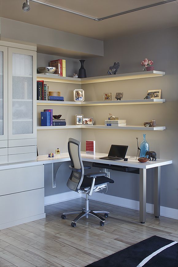 A white and silver home office corner desk is built-in to the end of the wall storage unit, extending the usable surface space. The open shelves above feature a few small books, family photos, a vase of flowers, some wild animal sculptures, and other collectibles. On the desk is a laptop, and the chair is a modern white, silver, and black office chair on wheels. Natural light streams in from the window just out of view at the right edge of the photo.