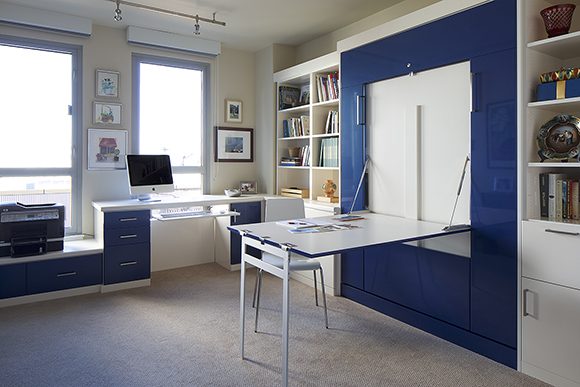 View of a San Francisco home office space with white drawers and silver hardware, on the right wall, with blue drawers and silver hardware on the left. To the right is a large matching blue Murphy bed or wall-bed in the closed position, with a dropdown white desk and chair. Open shelves appear either side of the wall bed cabinetry.