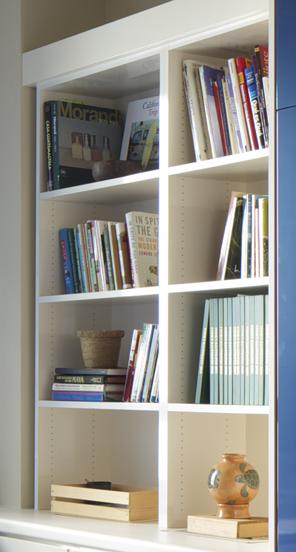 A closeup view of the far left set of open shelves, with books filling the top six cubbies, and a vase on top of a book on the bottom right, while the bottom left opening has a wooden slatted box containing office supplies.
