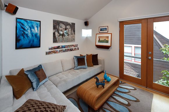 Looking further into the living room, a heavy wooden coffee table sits upon a blue, tan and brown patterned rug, in front of an L-shaped light-colored corner sofa with blue and brown throw pillows. Wall-mounted speakers save space and provide great sound for the party. Double glass doors surrounded by the same warm wood tones let in lots of natural light.