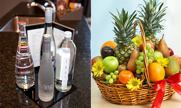 Two images side-by-side. Left image is of several luxury bottled waters on a quartz countertop. Right image is a basket of fresh fruit including pineapples, pears, apples, oranges, plums, tangerines, and mangos, plus fresh yellow flowers interspersed.