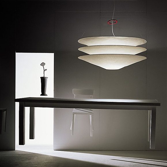 Three handmade shades are suspended one above the other by thin metal cables, while a single red iron ring provides a pop of color above. The paper shades of this pendant light called Floatation are crinkled, casting soft light over a dark dining table appearing to float in midair, with a white chair also floating behind, and a vase with a single rose, floating above the tabletop. Light is cast into the room from an open doorway at the back left of the image, creating interesting shadows across the floating pieces and the concrete flooring.