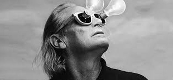 Photo of the famous lighting designer Ingo Maurer with shoulder-length hair, wearing a pair of sunglasses with lit lightbulbs coming out of the lenses. His head is turned up towards the sky.