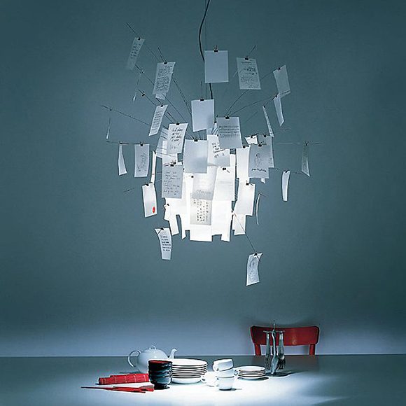 Photo of a pendant light created by a number of wire rods emanating out from the lighting fixture. On each rod is a piece of paper, some with writing on them, so that it looks like a giant round push-pin board of notes. Underneath is a red chair sitting behind a surface so that only the top rail is visible, and on the surface is a set of white dishes, a stack of black bowls, and some red cutlery.