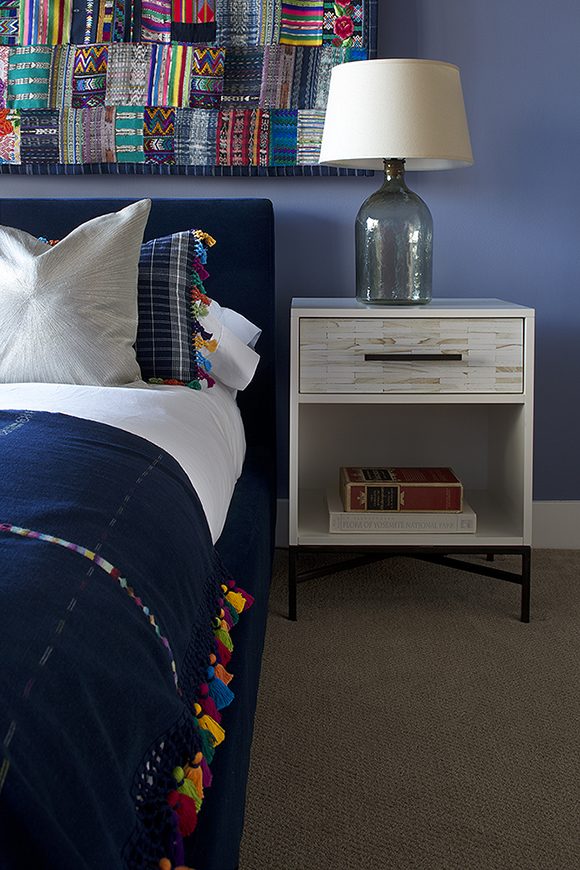 Close-up image of a bedroom, showing at the left, part of a bed with blue velvet headboard, white sheets, and blue pillow with multi-colored tassels, which echoes the multi-colored tassels on the blanket covering the bed. Above the headboard is a quilt made of many multi-colored and textured rectangles of fabric, sewn together. To the right, a light-colored bedside table with a single drawer hold two books underneath in an open space, and on top, a glass table lamp is topped by an off-white lampshade.