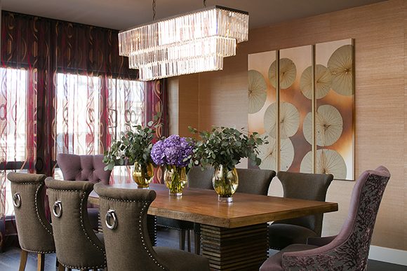 View into an open-plan dining area, featuring two contemporary purple head chairs with floral velvet backs, six brown velvet dining chairs with ring backs, and a dark wood dining table under a triple-tiered glass chandelier. Just beyond that, a gold and copper triptych of water lilies hangs over tan and brown textured wallpaper, with hot pink and orange patterned sheer curtains covering the triple window to the left.