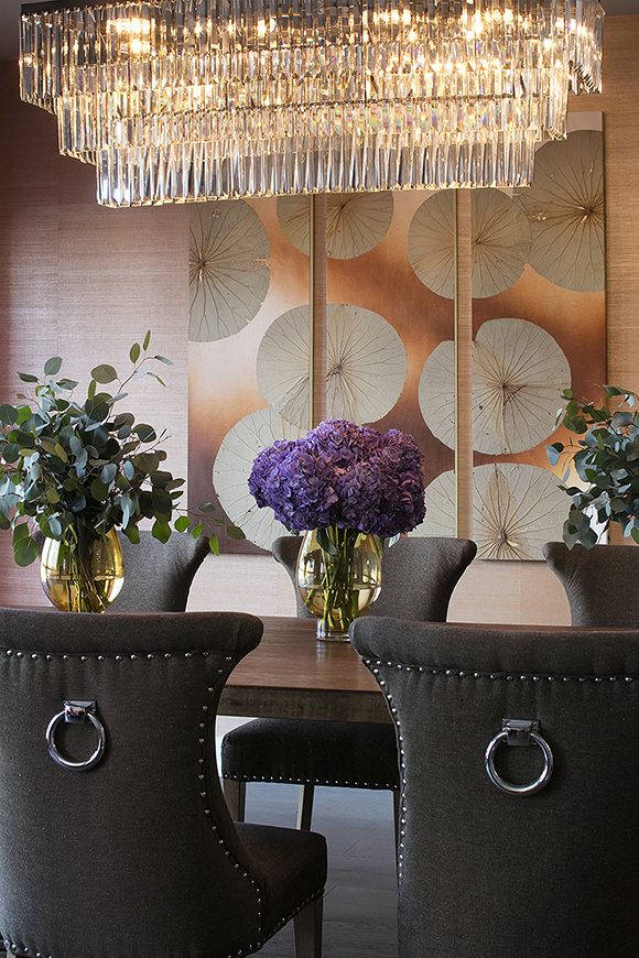 Close-up of a contemporary dining area, featuring brown velvet dining chairs with silver ring backs and silver nail heads around the edges, and a dark wood dining table under a triple-tiered glass chandelier. A vase of purple hydrangea flowers is in the center, with two vases of green cuttings either side. Behind, a gold and copper triptych of water lilies hangs over tan and brown textured wallpaper.