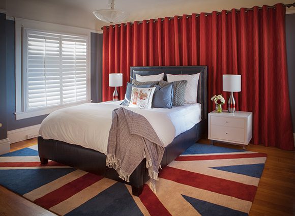 A large open spaced bedroom, with a huge Union Jack flag rug underneath a dark brown padded upholstered bed with white sheets, layered with white, grey, blue, and graphic pillows. White bedside tables have 2 drawers each, with bowling pin lamps and white shades. Behind the bed is a wall of bright red curtain. To the left is a window with slatted blinds in white, with white trim. The walls are painted a denim blue. Warm reddish wood flooring underneath the rug.