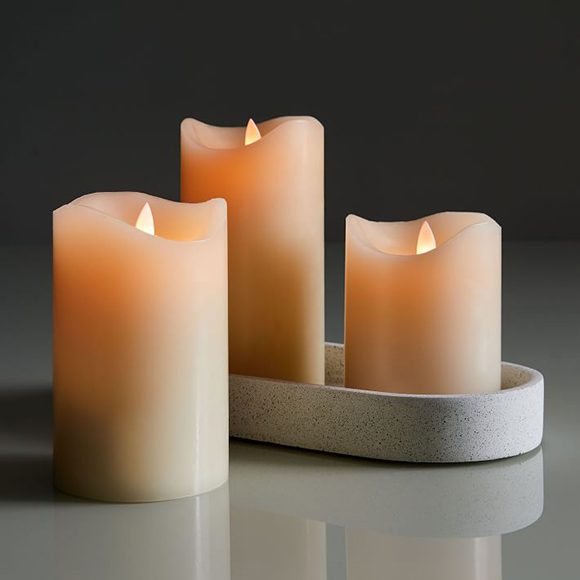 Three wax pillar candles in different heights, two in an oval container to protect the surface they rest upon, the third on a reflective grey countertop. All three candles are flameless, with lit faux flames peeking out at the top.