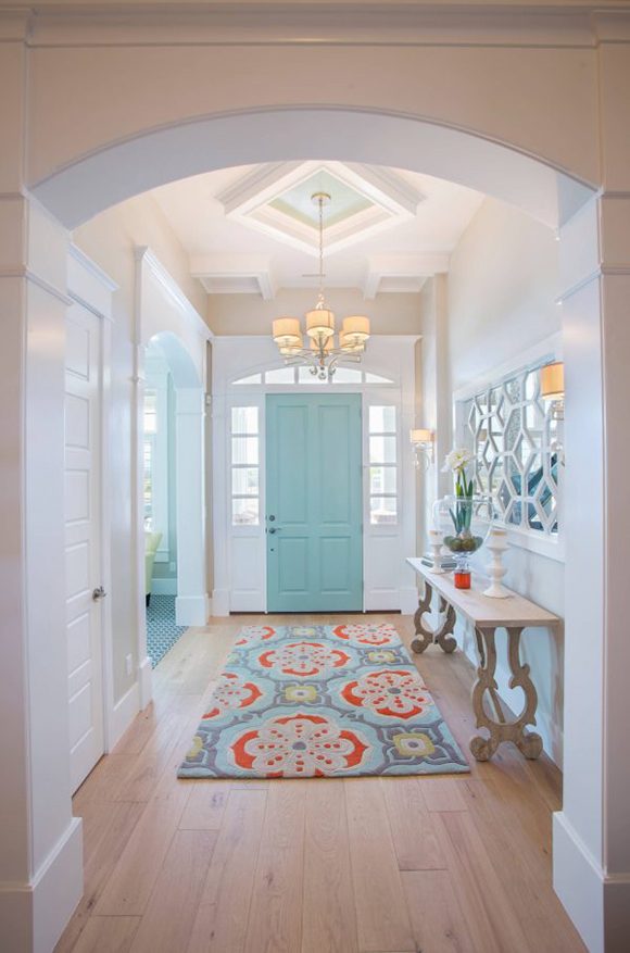 View down a hallway under an arched room separation, with white painted ceiling and doors and door trim, with the exception of the front door at the end, painted a robin's egg green-blue. A rug running along the hall echoes the door color and adds orange, yellow, and grey.