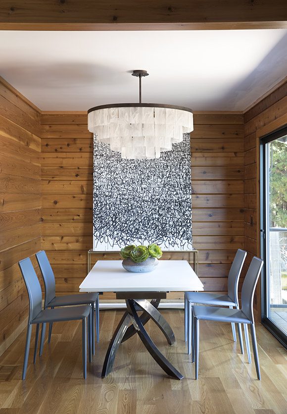 A contemporary condo's walls are clad in knotty cedar wood and accented with a white ceiling. A black and white canvas hangs on the wall behind the dining table, topped in white on dark twisty legs. Four modern slim leather chairs in a grey-blue surround the table. A tiered glass chandelier hangs above, ready to fill in when the light streaming in the sliding glass door at the right of the space is unavailable.