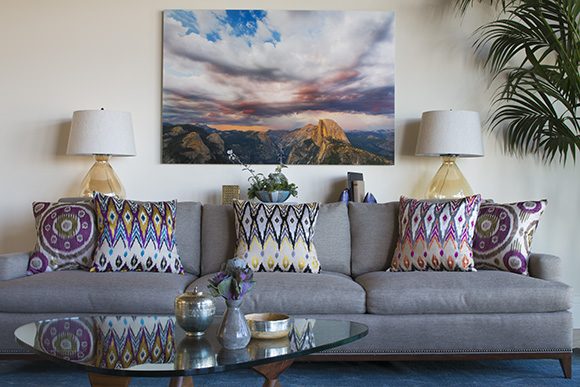 A living room vignette, showing a grey mid-century modern sofa with 5 multi-colored and multi-patterned throw pillows scattered across it. In front is a glass Noguchi-style coffee table, and behind the sofa are two yellow glass table lamps. Just above the sofa on the wall is a colorful photo printed on canvas of Yosemite Half-Dome.