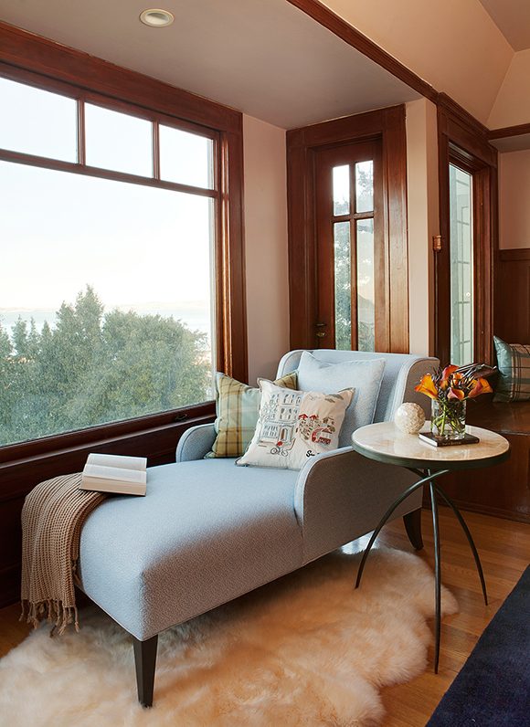 A custom-made light-blue chaise lounge sits atop a sheepskin rug, next to a triple-legged side table, dressed with several throw pillows, a tan blanket and an open book, in front of a large picture window without any window coverings, revealing a view of green trees.