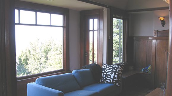 A living room view shows a bright blue sofa with dark blue patterned throw pillows, in front of a large Craftsman-style window, with green trees outside. The walls are half-covered in warm wood paneling.