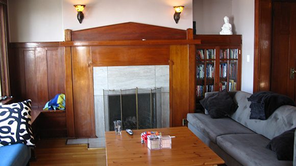 A Craftsman-style wood fireplace surround, with a built-in bookshelf to the right. To the left, wood paneling and a sliver of a bright blue sofa. To the right, a grey sofa with black pillows, in front of a large wooden coffee table. Two wall sconces are lit above the fireplace.