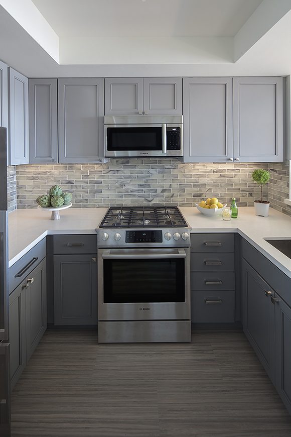 A modern compact U-shaped kitchen, with a silver and black range and oven at the center, and matching microwave above it. The top cabinets are a light grey with round silver handles, while the lower cabinets are a darker grey with horizontal silver drawer pulls and handles. The flooring is a multicolor horizontal pattern. The countertops are an off-white, with grey multishaded subway tiles as backsplash and running throughout the kitchen walls. The countertops are dressed with fruit and vegetables and bottled water in glass, plus a topiary-style plant.