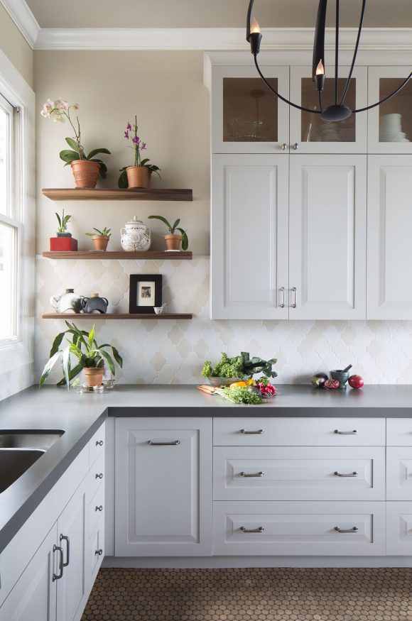 Left corner of a white farmhouse style kitchen showing open shelving with potted herbs and plants, a grey countertop over white cabinetry with silver drawer pulls, and a round metal pendant light.
