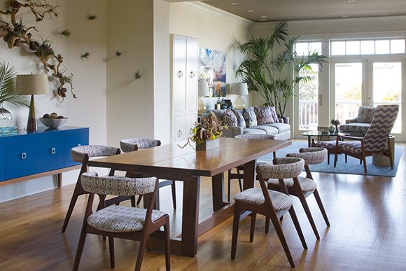 View from a dining area into an open plan living space. At the forefront is a mid-century modern wood table and wooden chairs with multicolor fabric seats and backs. Plants and natural driftwood sculptures appears at the back walls. A bright window wall and double door all made of glass stream light into the living space.