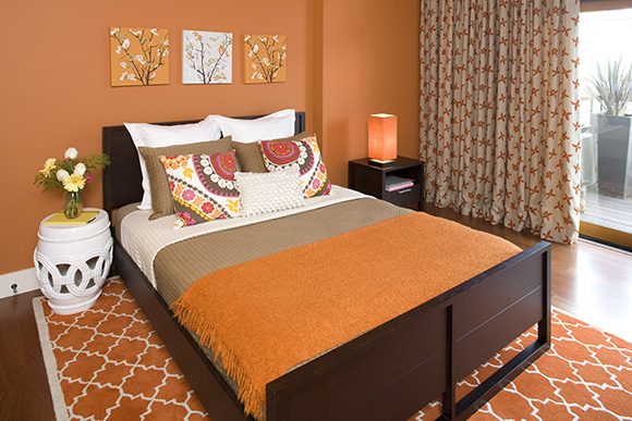 A tangerine and orange colored bedroom, with multiple patterns, textures, and several layers of pillows on the bed, plus tan blanket, white sheets, and orange comforter on a dark brown wood bed.