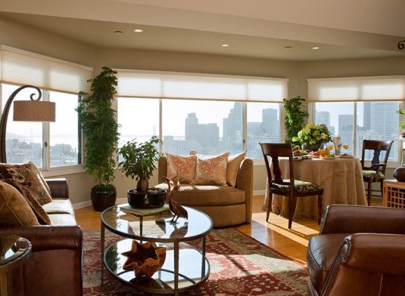 A traditional living and dining space in a high-rise condo, with a huge expanse of windows at the back, showing a city skyline. Multiple brown leather chairs and loveseats with throw pillows surround a glass and metal oval coffee table on top of a red-patterned oriental rug. To the right, a dining area is dressed with flowers and food and drink. Light streams in from the windows, hitting the blond wood flooring.