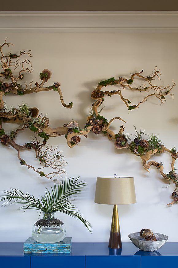 At bottom a bright blue console table supports a bronze lamp, silver bowl, and fresh fronds in a low vase, while above on the wall hangs a natural driftwood sculpture with airplants and moss.