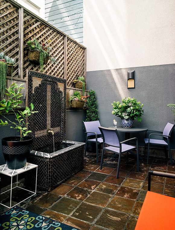 = A private interior courtyard in this Folsom Street San Francisco home looks like you’re actually somewhere in the Mediterranean or Morocco, with an intricately tiled water feature in front of a high wooden privacy fence, hung with multiple planters. The ground is covered in a coppery oversized tile, and a round table and blue chairs sits next to a plain grey wall, topped with a vase of flowers. In the foreground, an orange seat peaks through, while to the left we can just see a lime green, turquoise blue and brown geometric patterned indoor-outdoor rug.