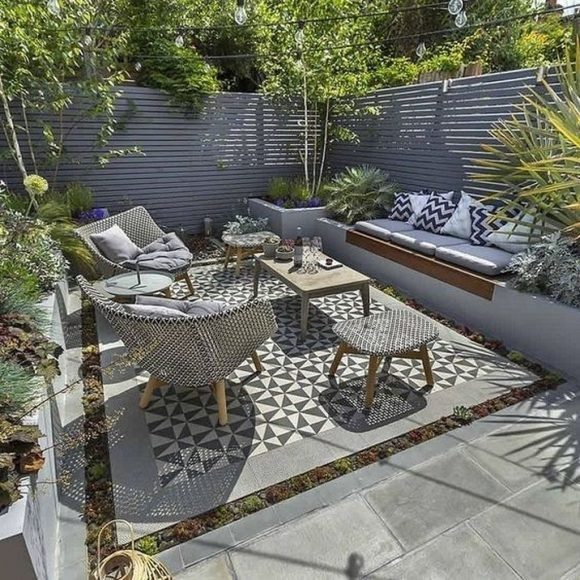 Grey slatted fencing form a corner of a yard with greenery above and behind. In front of the fending sits an outdoor living room, with grey woven chairs and footstools plus a grey bench seat with striped throw pillows surround a small table. Triangles form geometric patterns on the tiling underneath, forming a visual rug on a slab of concrete underfoot.