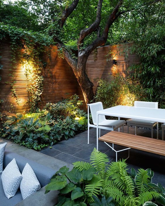 Warm reddish brown slatted wood fencing forms a corner behind a tree. Lights wash ivy tendrils hanging down the wall created by the fence, pooling in the green plants below. To the right is a modern, white rectangular table and two white chairs. A wooden bench seat is at the front, with more plantings in the center front. To the bottom right of the image is a grey outdoor sofa with throw pillows. Large grey slate tiles are both flooring and path underfoot.