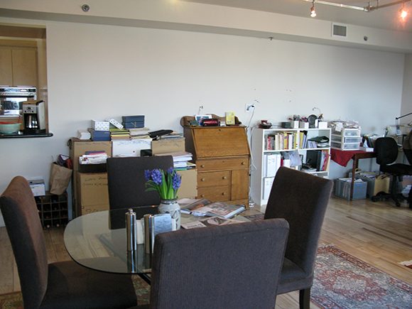 A 'before' image of the client's open-plan living/dining area shows multiple open shelves, a secretary desk and piles of boxes pushed up against the back wall, with a dining table towards the front. A passthrough to the kitchen is just visible at the left, and the rest of the white wall is blank.