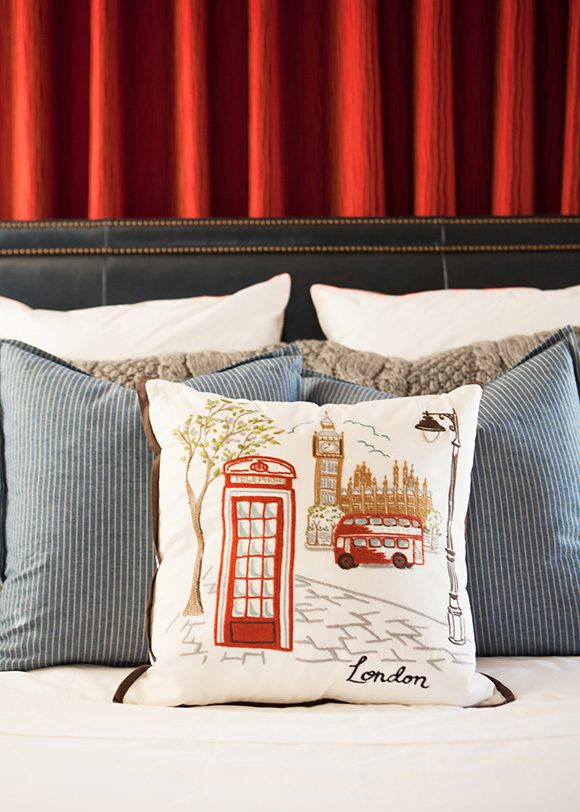 Closeup view of pillows layered on a bed. Behind the dark brown leather headboard with nailheads is a red curtain wall. The front pillow features graphics of London phone booth and bus, with two blue and white pinstriped pillows behind, and two textured beige pillows behind that, and finally two plain white pillows at the back.