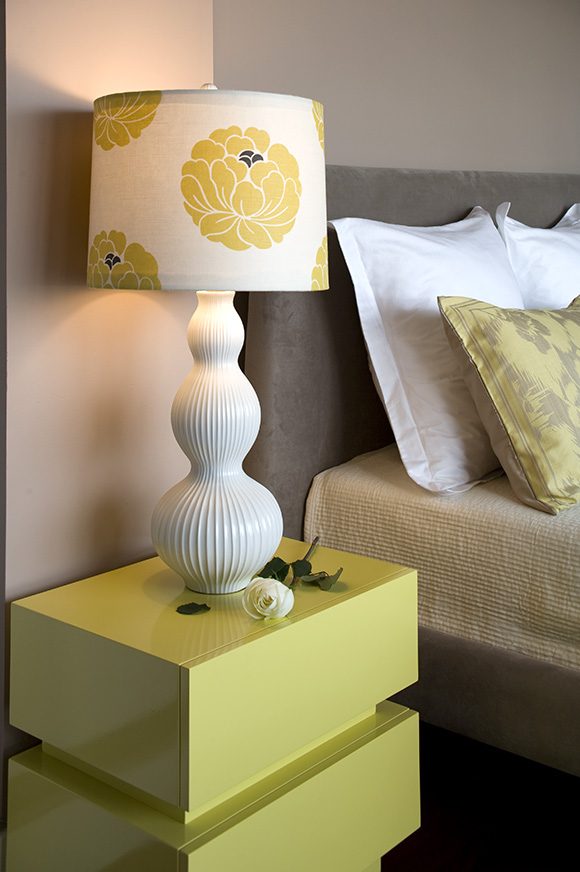 A bright yellow square melamine bedside table holds a white and yellow lamp next to a tan suede headboard and beige textured sheets with white pillows and a grey and yellow pillow just showing to the right.