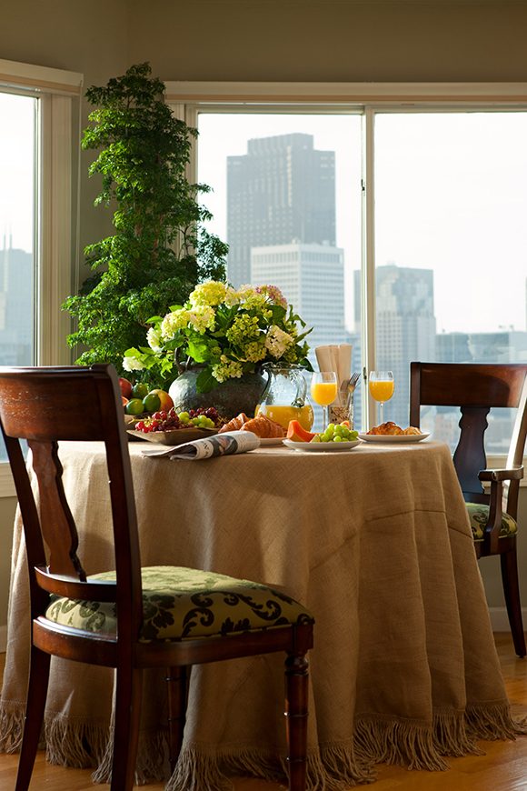A light-filled dining space shows a city view out the windows, while a traditional dining table draped with a light-colored tablecloth and two dark wood chairs sit atop warm wood floor planks. The table is dressed with flowers, orange juice and fruit for two, with a newspaper folded over.