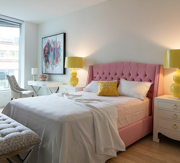 A girlie bedroom with white walls and a white desk and chair sits next to a bed with pink tufted headboard and white sheets and pillows plus a single yellow pillow, between two white bedside tables and yellow bubble lamps. Light streams in the through the window at the left.