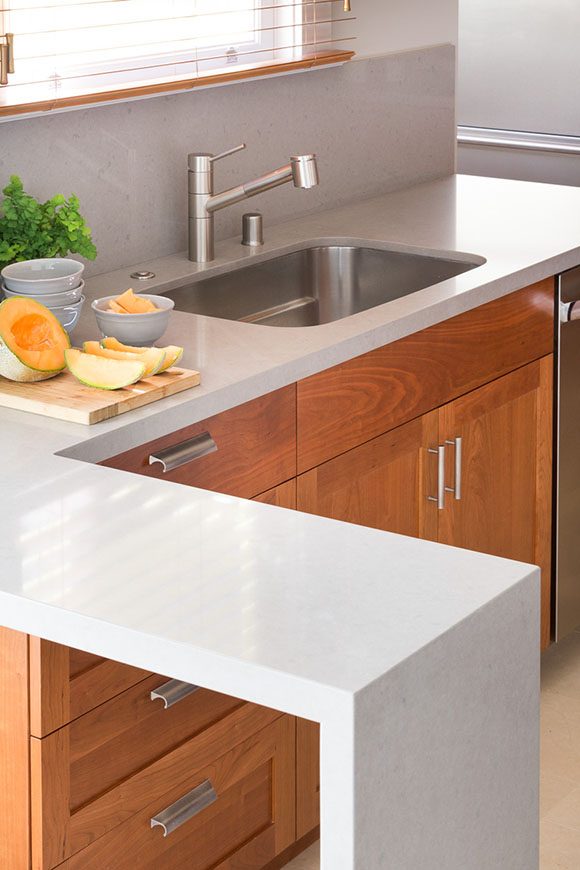 An L-shaped white counter surface continues around to the modern silver sink and faucet. The lower cabinets are cherry wood with silver drawer pulls and handles. Above the sink is a window with cherry wood blinds opens to allow light in. The counter is dressed with a cutting board of melon and several bowls waiting to serve.