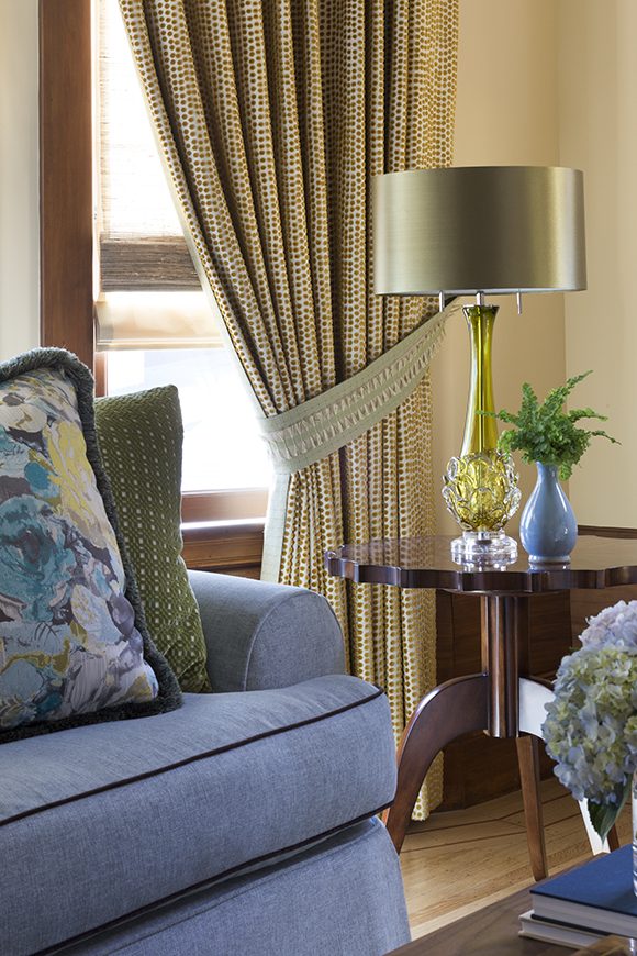 A vignette of a living room corner, showing a gold patterned drapery with a matching tie-back, a dark wood side table with a gold and glass lamp and small vase of greenery on it. Next to the table you see part of a periwinkle blue sofa with matching gold and floral pillows. A hint of a coffee table dressed with fresh hydrangea flowers on it just shows at the bottom right corner.