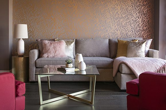 A living room vignette shows a pale violet three-seater sofa dressed with gold and pink fur pillows, and a pink cashmere blanket is thrown over one end. In front, a square glass table with gold cross-crossed legs is dressed with a small vase and books. To the left side, you see a small round gold side table and a white lamp. The wall behind the sofa is reflective patterns of gold plants scattered across a purple background color. In the forefront, two hot pink reclining chairs peek out at the left and right corners of your view.