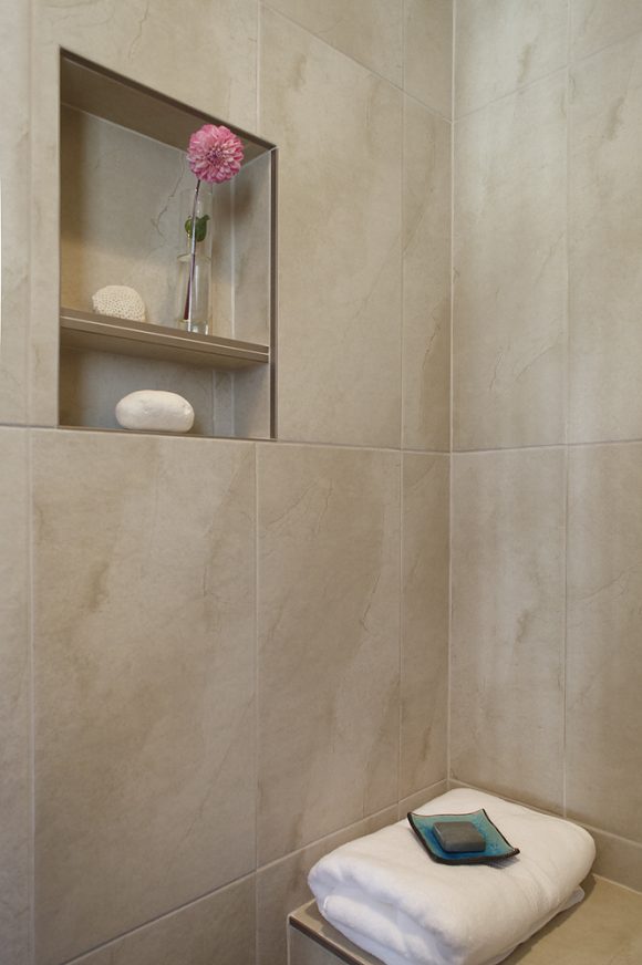 A beige marble and porcelain shower corner is dressed with a seat and white folded towel with a bar of soap. Above, an inset in the shower wall holds a shower scrub and a vase with a single pink flower.