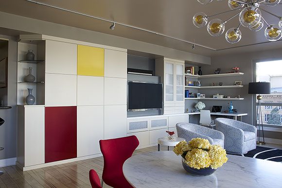 After the redesign, you see a large wall of custom storage, mostly in white with one yellow square cabinet door and one red rectangular cabinet door. A television and media storage is also built-in, with a corner built-in desk and open shelving at the far right. In front is a white marble table and red modern dining chairs, with a bubble pendant light above, and club chairs in the background.