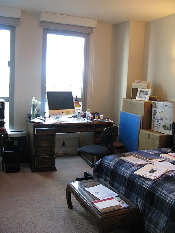 A bedroom with papers strewn all over the bed also has a desk with large Mac computer and keyboard, plus many boxes and shelves of folders, books and supplies pushed against the walls.